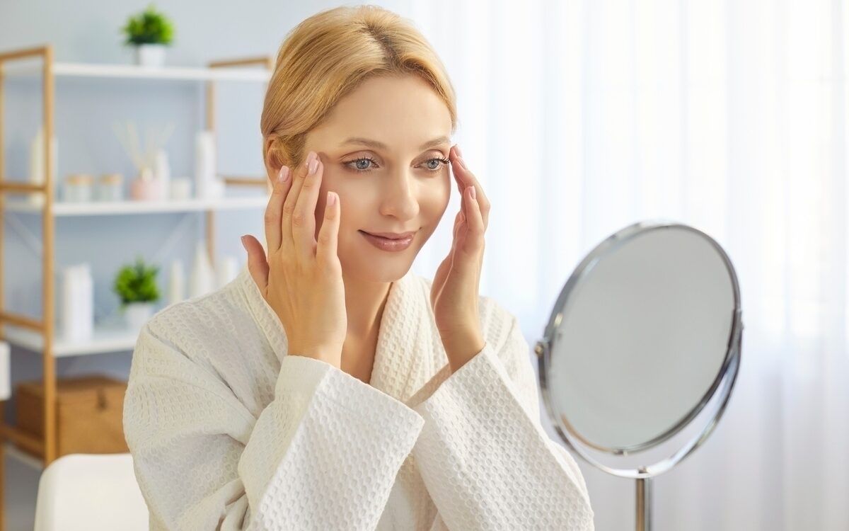 Woman gently touching her face while checking for uneven dermal fillers in a mirror