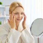 Woman gently touching her face while checking for uneven dermal fillers in a mirror